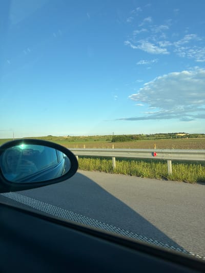 View from inside a vehicle moving along a road near Chirpan, Bulgaria. A side-view mirror is visible, reflecting a person holding a phone. A safety barrier and vegetation are adjacent to the road. The sky is blue with white clouds.