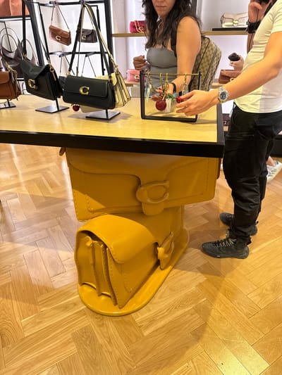 The image depicts the interior of a retail space in Sunrise, Florida, United States of America, likely a fashion accessories store. A person wearing a white t-shirt, dark trousers, and sneakers is standing near a display table. On the table are multiple handbags, including a large yellow bag prominently displayed, along with other handbags on a tiered display. A person with dark hair wearing a grey top is also visible near the table, apparently looking at a display stand with shiny keychains. The floor is wooden and arranged in a parquet pattern.