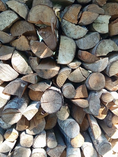 This media file displays a close-up, overhead view of a large, haphazardly stacked pile of chopped firewood. The logs vary in size and shape, with many showing the circular grain patterns characteristic of cut wood. Some pieces are split lengthwise, revealing the wood's texture and color, which ranges from pale beige to a warm brown with darker bark. The firewood is densely packed, creating an irregular surface with shadows and highlights cast by direct sunlight. The sunlight suggests it is daytime, and the overall lighting and shadows indicate a sunny day. There are no people or animals visible. The setting appears to be an outdoor space, likely a yard or storage area, given the presence of firewood. The location context provided is Ostrovtsy, Russia, which contextualizes the scene as a common sight in a region where firewood is used for heating. There is no visible text in the image.