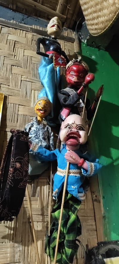 The scene in Cikanyere, Indonesia, depicts five traditional Indonesian wooden puppets, likely Wayang Golek, displayed vertically against a wall. The background consists of a woven bamboo matting wall on the left and center, transitioning to a solid green painted wall on the right.
From top to bottom:
1. An uppermost puppet with a pale, toothy face, a black torso, a light blue draped lower garment, and a red and gold adorned sash.
2. Adjacent and slightly below is a puppet with a striking red face, white eyes, a white headwrap, and a black shirt featuring a white checkered pattern. Its red-colored arms hold two thin wooden rods.
3. To the left, a puppet with a yellow-orange face, a mustache, and a smiling expression is dressed in a blue and white patterned long-sleeved shirt.
4. The largest puppet is positioned at the bottom, featuring a pale, exaggerated face with wide eyes, dark eyebrows, and a large open mouth revealing a long red tongue. It wears a light blue jacket with white trim, a red bracelet on its right wrist, and a green plaid lower garment. This puppet holds a long wooden rod vertically.
5. Partially visible on the bottom left is a dark fabric object with brown floral or batik patterns, alongside another wooden rod.
In the upper right background, a woven basket hangs. The scene is lit, suggesting an indoor environment, with slight shadows indicating an overhead light source.