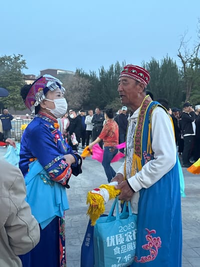 Two people in traditional Chinese clothing are standing outdoors. The woman on the left wears a blue dress with embroidered sleeves, a white face mask, and an elaborate silver headdress. The man on the right wears a red and white hat, a white shirt with a colorful vest, and holds a blue tote bag. Behind them, a group of people are gathered, some wearing masks, and others are dancing with colorful scarves. The location appears to be an outdoor plaza, with trees and buildings visible in the background. This is likely in Jinzhou, China, given the tote bag has Chinese writing on it and the people are wearing traditional Chinese clothing.