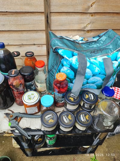 This is a high-angle, eye-level shot of a collection of jars, bottles, and a reusable shopping bag filled with what appears to be preserves or pickled items, arranged on a rough surface, possibly a tarp or old fabric, with some items placed on a black metal shelf. The scene is outdoors, with a weathered wooden fence in the background, suggesting a garden or backyard setting. The lighting indicates it's daytime, and the overall atmosphere is casual and utilitarian, possibly a collection of homemade goods or items being stored.
Prominently featured are several glass jars containing various colored preserved fruits and vegetables, some with visible spices like bay leaves. There are also tins of "Spichlerz Rusiecki" Polish culinary items. A large blue and white patterned reusable shopping bag, possibly containing more of the same, dominates the right side of the frame. A pair of gardening shears lies in the foreground, hinting at activities related to harvesting or preserving produce.
Visible text includes brand names like "Freshona" and "Spichlerz Rusiecki," along with some Ukrainian text on one of the jar labels. A timestamp "18.06.2026 12:53" is present in the bottom right corner. The scene does not contain any people, but the presence of the gardening shears suggests a person has recently been or will soon be involved in activities related to these items. The setting appears to be a domestic outdoor space, likely in Northampton, United Kingdom, given the provided context.
