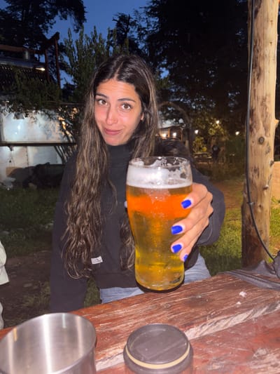 A brunette woman holds a glass of beer towards the camera, showcasing her blue fingernails. She is wearing a black sweatshirt and sitting at a wooden picnic table, where two metal containers are placed. The background features trees and an outdoor setting with lights. The photograph appears to be taken in the evening or at dusk.