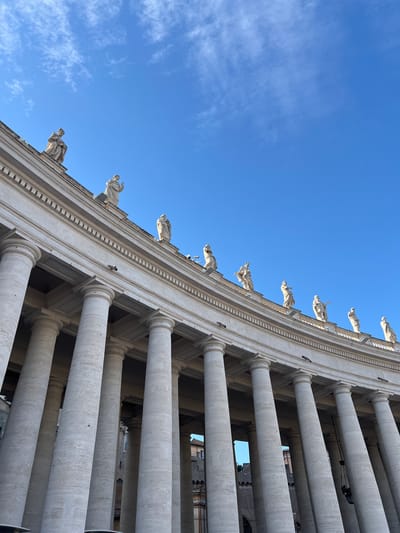 An extreme low-angle perspective captures a segment of the St. Peter's Square Colonnade in Città del Vaticano, Vatican City, under a clear, bright blue sky with sparse, wispy white clouds. The monumental colonnade, constructed from light-colored, textured stone (likely travertine), features multiple rows of large, cylindrical Doric columns, receding into the background. Above the column capitals and entablature, a series of at least ten human-sized statues, carved from similar light stone and depicting robed figures, stand along the roofline. The statues are evenly spaced and illuminated by strong natural sunlight, which highlights the architectural details and casts subtle shadows. Small, dark surveillance cameras or sensors are affixed to the upper facade of the colonnade at irregular intervals. Through the gaps between the columns on the right, dark, shadowed architectural elements of an adjacent structure are faintly visible. The colonnade curves gently from the lower left to the upper right, emphasizing its expansive scale.