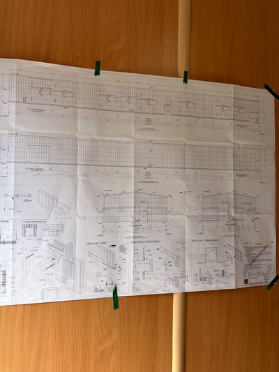 The image shows a large, unfurled architectural blueprint attached to a wooden wall with green tape. The blueprint is detailed, with grids, lines, text, and technical drawings. It appears to be a plan for a building, possibly a warehouse or industrial facility, as indicated by labels like "BRAMA" (gate) and "STREFA WZMOCNIONA" (reinforced zone). There are also sections labeled with numbers and possibly dimensions, along with various technical details and diagrams of building components. The overall impression is that of a construction project in progress, with the blueprint serving as a central document for planning and execution. The background is a plain wooden wall, suggesting an interior space, possibly an office or a temporary site office. The lighting is natural, coming from an unseen source, casting a soft light on the blueprint and wall. There are no people visible in the image. The location in the city of Stróża, Poland is mentioned, but there is no visual information within the image itself that directly indicates this specific location. The image is a static representation of the blueprint and its immediate surroundings.