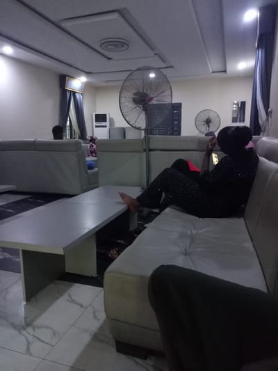 The image depicts an indoor lounge area, likely in Uke, Nigeria, characterized by a suspended ceiling with inset lighting and a fan. In the foreground, a long, light-colored coffee table is visible, with a beige sectional sofa extending to the right. A person, viewed from the back, is seated on the sofa, wearing dark clothing adorned with small white dots and using a mobile phone. Their feet are resting on the coffee table. To the left, another person with dark hair is seated on a separate part of the sofa, facing away from the camera. In the background, an air conditioner unit and another fan are present, along with curtains and a framed picture on the wall. The floor is tiled with a marble-like pattern.