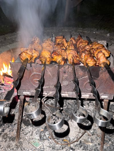 The image depicts several skewers of meat cooking over an open fire in what appears to be an outdoor setting. The meat is browned and appears to be seasoned, with some charring visible. Smoke is rising from the skewers, indicating they are actively cooking. The skewers are made of metal and have twisted handles. They are placed on a metal grill or brazier, which is situated on a bed of ash and embers. In the background, there are glimpses of greenery and what looks like a wooden structure, possibly a table or bench, suggesting an outdoor recreational area. A fire is visible to the left of the frame, providing heat for the cooking process. The overall scene evokes a sense of outdoor cooking and gathering.