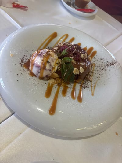 A dessert is presented on a white oval plate, positioned centrally on a white tablecloth. The dessert features a scoop of ice cream with swirls of purple and white, drizzled with caramel sauce. Adjacent to the ice cream is a dark, chocolate-based confection, possibly a lava cake or brownie, topped with chopped nuts, dried berries, and fresh mint leaves. The plate is dusted with cocoa powder and almond slivers, with further drizzles of caramel sauce artistically spread across the surface. In the background, slightly out of focus, a small white dish holds a shot of liquid, likely coffee or a liqueur, accompanied by a red packet. The scene suggests a restaurant setting, possibly in Urbanización Roquetas De Mar, Spain, given the context.