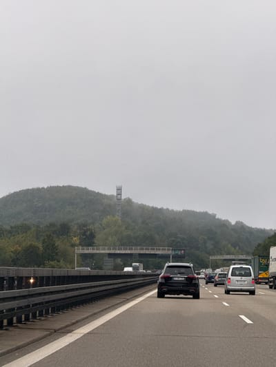 The image displays a multi-lane German autobahn, likely the A8 or A81, situated near Leonberg, Germany, on an overcast day. The perspective is from within a vehicle looking forward, capturing traffic.
In the foreground, the grey asphalt road surface features white solid and dashed lane markings. A metal guardrail with dark vertical posts runs along the left side of the highway.
The midground shows moderate to heavy vehicle traffic. A dark-colored SUV, identified as a Mercedes-Benz with the license plate "BB MB 2774" (Böblingen district), occupies one of the right lanes. To its right, a light-colored van, likely a Volkswagen, bears the license plate "LB C 2307" (Ludwigsburg district). Further ahead, a line of various vehicles, including cars and large trucks, is visible, extending into the distance. An elevated concrete overpass structure with traffic sign gantries spans perpendicular to the highway.
The background is dominated by a densely forested hill. A grey, lattice-style telecommunications tower stands prominently on the upper slope of this hill, partially obscured by atmospheric haze. The sky is uniformly grey and lacks distinct cloud formations. No individuals are explicitly visible, but their presence is inferred by the operation of the vehicles. The scene depicts vehicles in motion or stopped in traffic on a major highway.