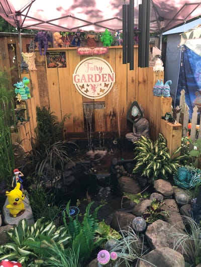 In the city of Severn, Canada, a backyard fairy garden is set up under a pink canopy tent. The garden features a small pond with a waterfall, surrounded by rocks and various plants. Several decorative items are placed throughout the garden, including figurines of mythical creatures like dragons and gargoyles, as well as pop culture characters such as Pikachu and Ash Ketchum from Pokémon. A statue of Buddha sits near the waterfall. Wind chimes hang from the tent's structure, and a sign reads "Fairy Garden." The overall scene is whimsical and elaborately decorated.
