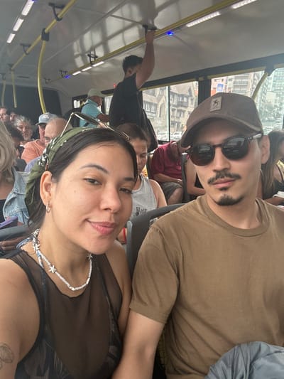 A crowded public transit bus in Mar del Plata, Argentina, serves as the setting for a selfie-style photograph. In the immediate foreground, an adult female with a light brown complexion, dark hair covered by a green patterned bandana, and silver hoop earrings, wears a dark sheer top and a white beaded necklace with starfish charms. She exhibits a faint smile directed at the camera. Beside her, an adult male with a light brown complexion, dark hair, mustache, and goatee, wears dark sunglasses, a brown baseball cap with an indistinct logo, and a plain brown t-shirt. He maintains a neutral expression, also looking directly at the camera.
Behind the primary subjects, numerous other passengers occupy the bus; one male is visible standing and gripping a yellow overhead rail. Another older male passenger in a light blue shirt and white hat is seated further back. The bus interior features yellow grab rails and overhead lighting. Through the bus window, a prominent multi-story building constructed of dark stone with ornate architectural details, consistent with historic chalets along Mar del Plata's coastal avenue (such as those in the vicinity of Avenida Patricio Peralta Ramos), is clearly visible. Parked vehicles and other urban structures line the street outside. The scene takes place during daylight hours.