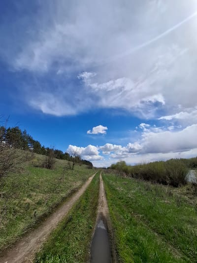 An outdoor landscape in Elets-Malanino, Russia, depicts a rural scene under a bright sky. The image is composed of a dirt track or path that stretches from the foreground into the distance, leading towards a horizon line. To the left of the path, a gentle slope of vibrant green grass is visible, dotted with various bushes and trees, some appearing deciduous with sparse branches, while others are lush with foliage.

On the right side of the path, a narrow body of water, possibly a ditch or small canal, runs parallel to the track. There are patches of what appears to be mud or standing water near the path, suggesting recent wet conditions or the nature of the terrain. The sky dominates a significant portion of the upper frame, showcasing a clear, bright blue expanse adorned with scattered white clouds, ranging from puffy cumulus formations to wispy cirrus clouds. A distinct lens flare from the sun is visible in the upper left quadrant of the sky, indicating a sunny day.

The time of day appears to be daytime, likely late morning or early afternoon, given the strong illumination. The weather is fair and clear. There are no people or animals visible, and no specific activity or event is taking place. The scene conveys a sense of openness, natural beauty, and tranquility. No visible text is present in the image.