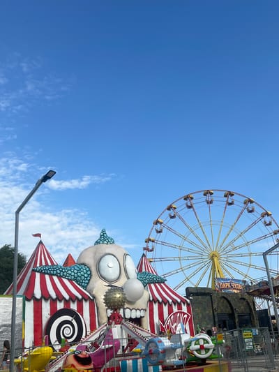 This is a vertical, medium-shot, outdoor photograph taken on a clear day with a blue sky and scattered white clouds. The scene depicts an amusement park in Golubitskaya, Russia, dominated by a large, stylized clown face structure with a gaping mouth, wide eyes, and a bumpy, teal-colored headpiece. The clown is part of a carnival-like setup with red and white striped tents and a ride featuring small, colorful vehicles. In the background, a large Ferris wheel with yellow gondolas is visible, along with a sign that reads "ПАРК ЧУДЕС" (Park of Wonders). A street lamp is present on the left side of the frame. The overall impression is of a vibrant and somewhat unsettling amusement park attraction.
