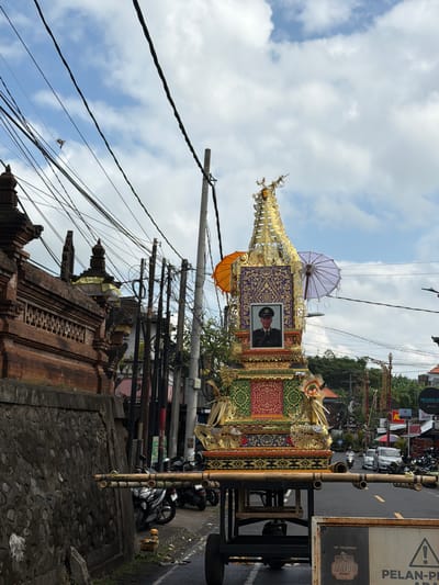 This image captures a traditional Balinese cremation ceremony (Ngaben). The central subject is a large, ornately decorated wooden effigy, possibly a sarcophagus or a representation of the deceased, mounted on a cart. The effigy is gilded with intricate patterns and features a portrait of a man in uniform, likely the person being cremated. White and orange umbrellas are displayed on the effigy, common elements in Balinese processions.

The scene is set on a street, indicated by the asphalt road, traffic signs, and parked motorcycles. The background shows buildings and lush greenery, suggesting a semi-urban or village setting. Overhead, a complex network of power lines crisscrosses the sky. The sky is partly cloudy, with patches of blue visible, indicating daytime.

The activity taking place is a funeral procession. The effigy, carried on the cart, is being moved through the streets, likely towards the cremation site. The presence of motorcycles and people in the background, though partially obscured, suggests a procession with participants. The overall atmosphere appears solemn yet ceremonial, as is typical for such events.

Notable details include the highly detailed craftsmanship of the effigy, the vibrant colors of its decoration, and the dense arrangement of electrical wires. A sign with a yellow triangle warning symbol and the text "PELAN-PELAN" (meaning "slowly" in Indonesian) is visible on the right. Another sign reads "MAYOR KOPI". The location context of Klungkung, Indonesia, strongly aligns with the cultural elements depicted.