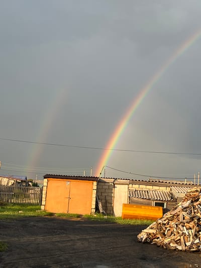 A double rainbow arcs across a grey, cloudy sky over a rural scene. In the foreground, a pile of split firewood is stacked on dark, soiled ground. Behind the firewood is a building with a corrugated metal roof. To the left of this building is another structure with large, orange double doors and a corrugated metal roof. Further left, a wooden fence encloses a grassy area with a small building with a blue roof. The location is described as Тимирязев ауылдық округі, Kazakhstan.