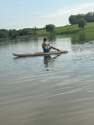The image shows a person balancing on a paddleboard in a body of water, likely a lake or wide river, in Novosinkovo, Russia. The individual, who appears to be female, is in a partially upright position, with one leg extended. The paddleboard is white and yellow, and the words "GO BOARD" are visible on its surface. The water's surface is rippled, reflecting the surrounding trees and sky. The background includes green fields and trees along the shoreline. A power line can be seen on the left side of the image.