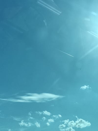 The image is a photograph of a bright blue sky with scattered clouds. A thin, white contrail from an airplane stretches diagonally across the upper middle portion of the frame. Towards the right side of the frame, a faint lens flare is visible. In the lower part of the image, several fluffy white clouds are dispersed. The overall scene suggests an aerial view or a clear day with an airplane passing overhead. The location is Divišov, Czechia.