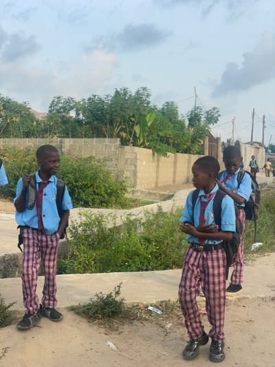 The image depicts an outdoor scene in Mosafejaw, Nigeria, featuring three young male individuals, identified as students by their uniforms, standing on a path under an overcast sky.
The uniforms consist of light blue short-sleeved shirts, maroon and white striped ties, red and white plaid trousers, and dark closed-toe shoes. Each student wears a black backpack. The individual on the far left stands facing right, his left hand in his trouser pocket, looking towards the central figure. The central individual stands facing left, arms crossed over his chest, wearing a gold-buckled belt, and looking towards the left individual. The third student is positioned behind and to the right of the central figure, partially obscured, also facing left. Several indistinct figures are visible in the far background on a dirt path to the right.
The immediate ground transitions from a concrete slab on the left to a dirt path on the right. Green bushes line both sides of the path. A grey concrete block wall extends across the midground, behind which abundant green vegetation, including tall, slender trees and broad-leafed plants consistent with banana or plantain trees, is visible. A dark metal gate is discernible behind the wall on the right. Utility poles with overhead wires are present in the far right background. White debris, possibly paper or plastic, lies on the dirt path in the foreground. The sky is largely covered by grey and white clouds.