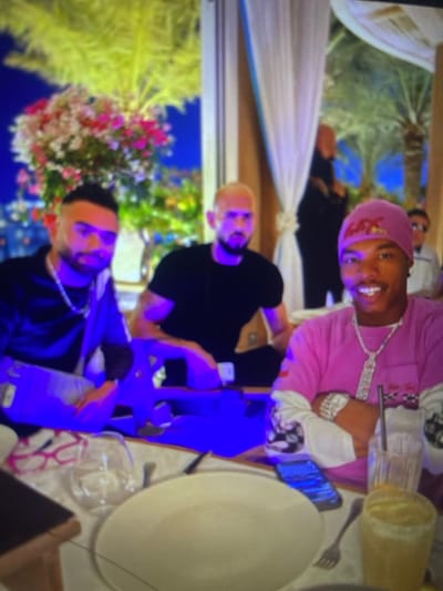 Three men are seated at a table. The man at the top wears a pink outfit with a matching head covering. Below him is a man in a black shirt. The bottom man wears a dark jacket with a silver chain. A white plate, silverware, and drinking glasses are arranged on the table. The scene is brightly lit, possibly outdoors. There are white draperies and a floral arrangement behind the men. Two phones appear to be on the table.