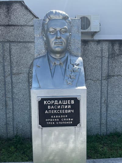 A silver bust of a man, identified by an engraved plaque as Kordashev Vasily Alekseevich, stands prominently outdoors. The bust depicts the man from the chest up, wearing a suit and tie, adorned with several star-shaped medals on his left lapel. The plaque below the bust reads "КОРДАШЕВ ВАСИЛИЙ АЛЕКСЕЕВИЧ КАВАЛЕР ОРДЕНА СЛАВЫ ТРЕХ СТЕПЕНЕЙ". The bust is mounted on a grey pedestal. Behind the bust, a rough-textured grey wall is visible, with vertical lines suggesting construction or a natural pattern. An air conditioning unit is mounted high on the wall to the right of the bust. Green grass and weeds grow at the base of the pedestal and wall. The scene appears to be a memorial or monument. The image is likely taken in the city of Takhtamukai, Russia, where this monument is located.