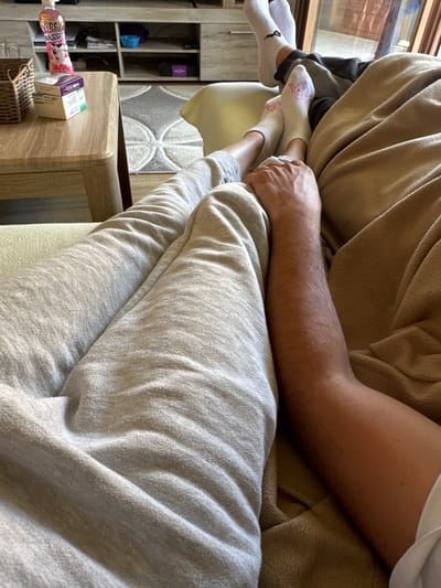 Two individuals are lying on a sofa, their legs and arms visible. One individual's arm, with visible arm hair, rests on a tan blanket. The other individual's legs are clad in grey sweatpants, and their feet are wearing white socks, one with a pink design. The scene appears to be indoors, with a coffee table to the left holding a basket and boxes. In the background, a shelf unit is visible with various items on it, and a rug is on the floor. The lighting suggests it is daytime. There is no indication of the city of Augsburg, Germany.