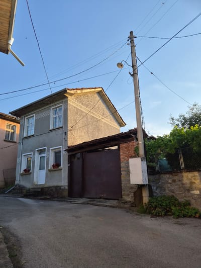 The image depicts a street scene in the city of Kaleytsa, Bulgaria. The focal point is a two-story gray house with white-trimmed windows and a white door. The house has decorative triangular patterns above the windows. A large, dark metal gate is visible to the right of the house, partially obscured by a brick structure. A telephone pole with a street lamp and various wires stands prominently in the foreground. To the right of the pole is a white electrical box. The street is paved with asphalt and appears to be wet in some areas. A stone wall and a chain-link fence with lush green vegetation are visible behind the gate and telephone pole. The sky is a clear blue with a few small birds flying. The lighting suggests it is either early morning or late afternoon.