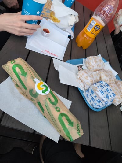 The image depicts a casual outdoor dining scene at a Jari-Pekka service station in Hartola, Finland. A dark wooden slatted table is covered with various food and drink items. On the left, a person's left hand grasps a light blue paper cup, marked "0.5 L" and bearing white star graphics and Finnish text "OLEN SUOMESSA VALMISTETTU." Next to the cup, a small white plastic cup contains red ketchup. Centrally positioned is a 1.5-liter clear plastic bottle of Jaffa brand orange soda, featuring a yellow label with a barcode and recycling symbols. Behind the Jaffa bottle, a paper container holds french fries. In the foreground, a sandwich from Subway is wrapped in its characteristic brown paper with green arrow patterns and a circular Subway logo sticker, resting on a white paper napkin. To the right, a blue plastic tray holds six individually wrapped food items resembling small burgers or sliders; five are wrapped in light brown paper with a repeating 'H' and star pattern, while one is wrapped in blue paper with the same pattern. A person's right hand is visible on the far right, holding one of these wrapped food items. White paper napkins and other paper wrappers, one bearing the "JARI-PERKKA www.jari-pekka.fi" logo in red, are present on the table. A red surface is partially visible in the background, to the right of the Jaffa bottle. Part of a person's leg is visible beneath the table on the lower right.