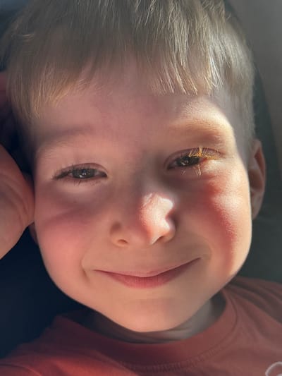 In Vykhodnoi, Russia, a close-up, eye-level shot captures a young boy with fair hair and fair skin. The boy is looking directly at the camera, his head tilted slightly and his left hand resting on his cheek. His eyes are open, with the right eye appearing more prominent due to lighting. The boy's lips are parted in a gentle smile. A strand of hair falls across his forehead, and sunlight illuminates his face, highlighting his features and casting shadows. He is wearing a rust-colored t-shirt. The background is dark and indistinct, suggesting an indoor or shaded outdoor setting.