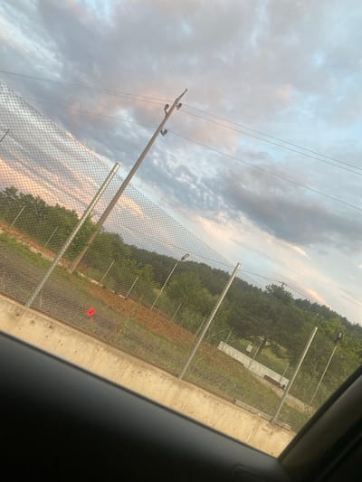 The image depicts a view through a car window of a chain-link fence, with a concrete barrier in the foreground. Beyond the fence, there are trees, a dirt area with some orange markers, and a white structure. Overhead, there are power lines and a cloudy sky with hints of pink and blue. The scene is outdoors, suggesting a roadside or perimeter setting. There are no people or animals visible in the frame. The location is specified as Ablanitsa, Bulgaria.