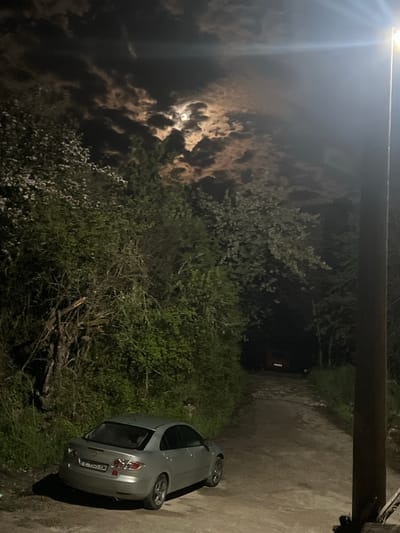 The image, taken at night in Kribul, Bulgaria, depicts an outdoor scene dominated by natural elements and human infrastructure. A light grey or silver Mazda 3 hatchback is parked on the right side of a concrete or asphalt path, which recedes into the darkened background. The car's license plate, "E 7305 BM," is visible on its rear.

The path is bordered on the left by dense green foliage, bushes, and trees, some of which display light-colored blossoms. In the night sky above the vegetation, a bright, full moon shines through dramatic, patchy clouds. To the far upper left, a powerful artificial light source, likely a streetlamp, casts a strong glare and visible light beams, illuminating a portion of the sky. A dark, thick utility pole or beam extends horizontally across the top of the frame. In the distance, at the end of the path, a faint red object with a vertical rectangular reflector or light can be discerned.

The scene is tranquil, with no human activity explicitly depicted. The time of day is clearly night, characterized by the prominent moon and the reliance on artificial lighting. The combination of moonlight, artificial light, and deep shadows creates a high-contrast and atmospheric setting.