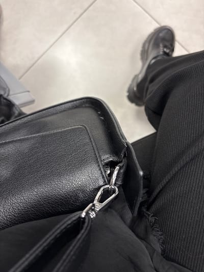 A high-angle, top-down shot captures the lower portion of a person's body and a bag, set against an indoor tiled floor. The foreground is dominated by a black, textured bag, appearing to be faux leather, with a prominent silver-toned metal swivel snap hook. A seam and what appears to be an outer pocket or flap are visible on the bag's surface. To the right, a person's right leg is clad in black trousers, featuring a visible ribbed or corduroy-like texture and a distressed or torn hem near the bag. Further back, the person's right foot is encased in a black, chunky-soled boot with visible laces and a thick, ridged sole. The floor is composed of light-colored, possibly off-white or light grey tiles, with distinct grout lines forming a geometric pattern. A bright reflection or light source illuminates a section of the floor in the upper right. A blurry, light-toned object is partially visible in the upper left corner. This scene is located in the city of Bol'shie Kabany, Russia.