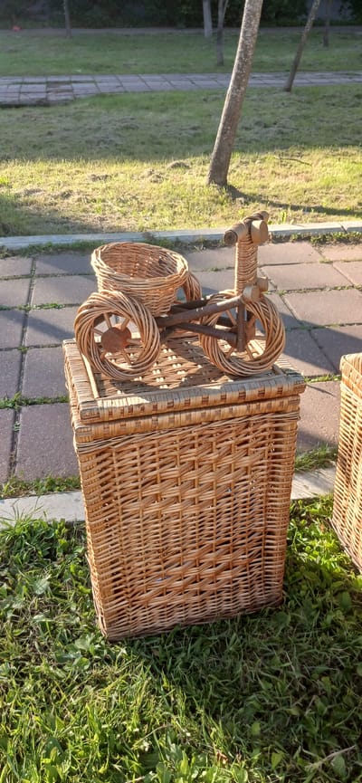 This media file shows a wicker picnic basket with a detached wheeled stand. The basket is made of natural woven material and has a rectangular shape with a lid. The wheeled stand is also constructed of wicker and features two circular wheels and a handle. The items are placed outdoors on a paved path with grass and foliage surrounding them. A tree trunk is visible in the background, suggesting a park or garden setting. The lighting indicates it is daytime, with sunlight casting shadows. There is no visible text in the image. The overall scene conveys a sense of leisure or preparation for an outdoor activity.