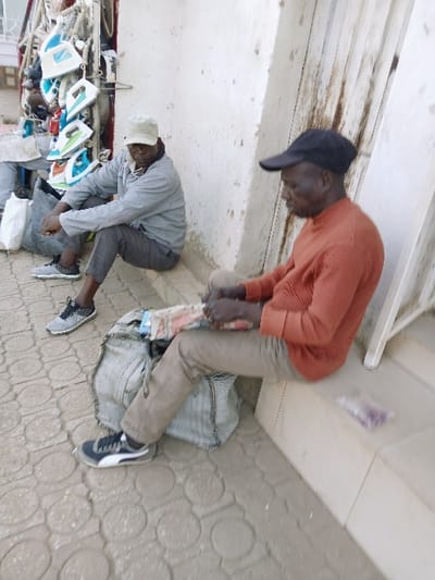 The image depicts an outdoor scene at Kungulu School, Zambia, featuring two adult males seated on a raised concrete ledge next to a building.
On the left, an individual is wearing a light-colored cap, a grey long-sleeved top, dark grey trousers, and grey and black sneakers. His hands are clasped in front of him. A large dark fabric bag rests next to his left leg, and a white plastic bag is visible further left. Behind this individual and to his left, multiple electric irons in various colors (primarily white, blue, and orange) are vertically displayed on a rack.
On the right, a second individual wears a dark baseball cap, an orange long-sleeved knitted shirt, light brown trousers, and black and white sneakers. He is holding small items in his hands over a patterned fabric item resting on his lap. A large dark fabric bag rests on the paved ground between his legs. A small translucent purple plastic bag containing unspecified items lies on the ground to his right.
The ground is paved with light grey interlocking concrete pavers embossed with a circular pattern. The building wall behind the men is light-colored and exhibits surface discoloration and weathering.