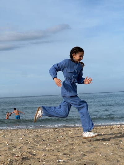 The image depicts a young woman in a blue jumpsuit and white sneakers appearing to levitate over a beach in M'Diq, Morocco. Her body is oriented horizontally, with her right foot hovering slightly above the sand. Her arms are extended outward as if balancing. In the background, the ocean extends to the horizon. Two figures are partially submerged in the water near the shoreline. The sand is light brown, containing small shells. The sky is blue. The image is rotated clockwise.