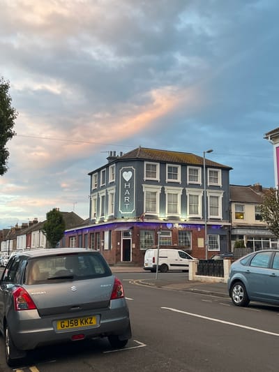 The image captures a street scene in Eastbourne, United Kingdom. The main subject is a three-story building with a sign that reads "THE HART" above the door, indicating it is a pub or similar establishment. The building's façade is primarily gray with white window frames and decorative elements. The sign features a heart symbol with the text "HART" inside. A vehicle with a license plate "GJ58 KKZ" is parked in the foreground, and a few other cars are visible further down the street. The sky is filled with soft clouds, tinged with hues of pink and blue. Additional buildings and a few trees line the street. The image shows a quiet urban setting.
