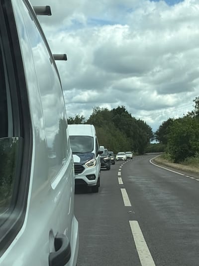 The image depicts a Google Street View car driving on a winding road in Flitton, United Kingdom. The road is asphalt with white line markings. The Street View car is white and has a camera apparatus mounted on top. The sky is overcast with thick clouds. The environment includes trees and vegetation lining the road. The image is taken from inside another vehicle, showing a partial view of its roof and a side window.