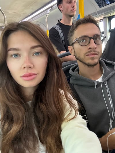 A selfie taken by a woman with long, wavy brown hair and light blue eyes, presumably on a public transit vehicle in Sdot Dan Regional Council, Israel. She is wearing a light-colored top. Behind her, a man with glasses and a light beard is visible; he is wearing a gray hoodie. Another man is partially visible in the background to the left, wearing a striped shirt. The interior of the transit vehicle includes handrails and windows.