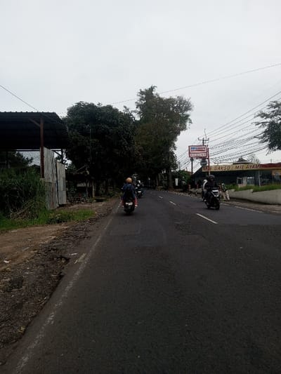 The image displays an overcast street scene in the city of Sat Thay (East), Myanmar. A two-lane asphalt road, dark in color with visible patches and cracks, recedes into the background under a uniformly grey sky. White dashed lines mark the lanes, and a solid white line defines the left edge of the paved surface. The unpaved left shoulder is uneven, displaying potholes, dirt, and overgrown green grass and weeds. On the far left, a structure with a dark corrugated metal roof supported by wooden posts and partial corrugated metal walls is visible. Dense, dark green trees line the road on the left. On the right side, a low, light-colored concrete barrier separates the road from commercial buildings. A red and white "Alfamart" sign is prominent, positioned above a sign advertising "MIE BAKSO & MIE AYAM." Numerous utility wires are suspended overhead across the right portion of the frame. Four motorcycles, each with a single rider, are visible on the road, traveling away from the viewer. The closest rider wears a light-colored helmet and dark clothing, carrying a backpack. A pedestrian in light attire is walking on the far right, near the commercial storefronts. No direct interactions between individuals are discernible. The scene is illuminated by diffused light consistent with an overcast sky.