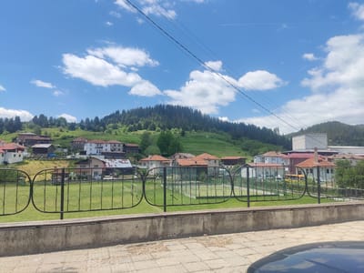 The image shows a view of Borino, Bulgaria, featuring a scene with a decorative metal fence in the foreground. Behind the fence is a grassy area, possibly a sports field or park. Beyond this area is a village setting with houses having red tile roofs. A forested hill is visible in the background, under a blue sky with scattered white clouds. Power lines stretch across the upper part of the frame. A concrete bench sits in front of the fence. The corner of a car is visible in the lower right of the frame. The time of day seems to be midday with natural light.