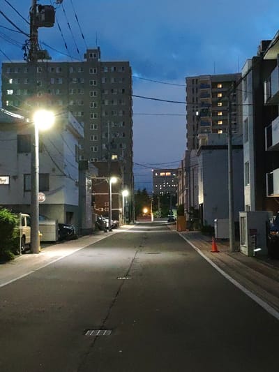 The image depicts a street scene in Sapporo, Japan, at dusk or early night. The street is paved and shows markings for a white line on the right side. Several buildings line the street, including multi-story apartment buildings with illuminated windows, indicating occupancy. Utility poles with numerous wires are visible, a common sight in urban Japanese settings. Streetlights illuminate the road and sidewalks. A white sign with Japanese characters, likely a warning or informational sign, is attached to a pole on the left. Cars are parked along the sidewalk on both sides of the street. An orange traffic cone is placed on the right side of the road. The sky is a deep blue with some clouds, suggesting the transition from daylight to darkness. The overall atmosphere is quiet and urban.