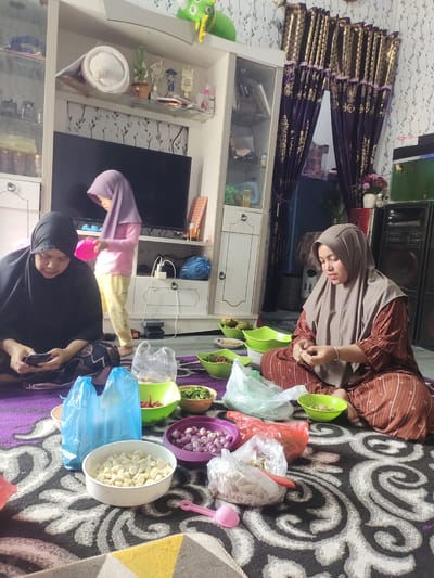 Two women and a young girl are sitting on a patterned rug in a living room, preparing food. The woman on the left, wearing a black hijab and dress, is looking at her phone. The woman on the right, wearing a beige hijab and a brown patterned dress, is focused on peeling green beans. The young girl, in a purple hijab and yellow patterned pants, stands behind the woman on the left, holding a pink bowl.
Several bowls of chopped ingredients are spread around them, including garlic, shallots, and chilies. There are also plastic bags filled with food. The setting appears to be a home in Simpangantara, Indonesia, with a television, shelves filled with decorative items and trophies, and curtains visible in the background. The lighting suggests it could be daytime. The overall atmosphere appears to be one of a shared activity, possibly for a meal. No visible text is present.