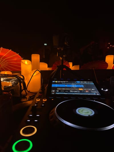 The image depicts a dimly lit, atmospheric scene, likely at a night event or performance, with a DJ setup in the foreground. Several large, illuminated LED candles of varying heights stand prominently, casting a warm glow. To the left, a clear plastic container with some items inside is visible. In the background, several red paper parasols are arranged, adding a touch of decorative color to the dark environment. A DJ mixer with a screen displaying waveforms and controls is central to the foreground, with illuminated buttons and a spinning platter. The overall ambiance suggests a relaxed, possibly musical, gathering. The location in Ban Bang Thao, Thailand, is not visually evident from the image itself. There are no discernable people or direct interactions captured in the frame.