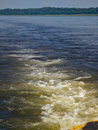 A wide shot captures a turbulent expanse of dark blue water, leading to a frothy, golden-brown wake in the foreground. The wake, characterized by swirling foam and disturbed water, suggests movement, possibly from a vessel. In the distance, a dense, green treeline stretches across the horizon under a pale, overcast sky. The scene appears to be a natural body of water, such as a river or lake, with no visible structures or individuals. The location is the Pakhra River in the village of Pogodaevo, Russia.