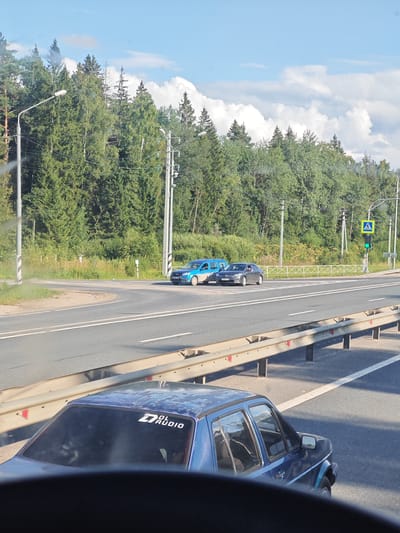 A blue minivan and a dark sedan are stopped at a traffic light on a two-lane road. The road curves to the right, with a tree line and a clear sky in the background. The minivan is in the left lane, and the sedan is in the right lane. The traffic light is green for the vehicles. A pedestrian crossing sign is visible next to the traffic light. The scene appears to be on the outskirts of Torzhok, Russia, given the presence of trees, a relatively quiet road, and typical Russian road infrastructure.
