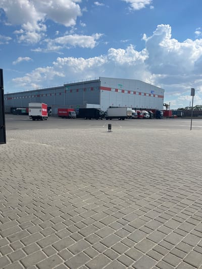 The image depicts a bright, sunny day with fluffy white clouds in a clear blue sky. The scene is an outdoor paved area, likely a logistics or distribution center, with a large, modern building in the background. The building is light gray with red and white accents and has numerous windows. There is visible text on the building which includes "УКРПОШТА 3" and "НОВА ПОШТА".

A row of trucks is parked along the side of the building. Some of these trucks have "НОВА ПОШТА" printed on their sides in red lettering. The pavement is made of interlocking gray bricks. A lamppost stands tall in the distance, and the overall impression is one of a functional, operational industrial or commercial space. The image is taken from a slightly elevated, tilted perspective. There are no people visible in the image. The time of day appears to be midday, given the strong sunlight and clear sky.