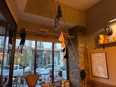 The image depicts the interior of a commercial establishment, likely a restaurant or bar, situated in Orléans, France, with a view through large windows onto a city street.
**Environment and Decor:**
From the ceiling, several thick ropes of natural fiber descend, adorned with various items of clothing, including one black bra, two white bras, one light blue/grey bra, and multiple grey and black fabric pieces, possibly shirts or rags. A light-colored wooden lattice structure is visible on the ceiling. A prominent corner column or wall section is faced with a multi-toned stacked stone veneer. An adjacent wall is painted a plain, light taupe color. Mounted on this wall is a single wooden shelf holding a yellow cylindrical tin labeled "BISOT," a small red box, a yellow toy vehicle resembling a taxi, and a dark circular object featuring a gold compass-like symbol. Below the shelf, a framed map titled "CARTE DES VINS FRANÇAIS" is displayed. In the immediate foreground, a table corner is visible, set with multiple wine glasses and a vintage-style red counter scale with a gold pan. Partially visible at the bottom of the frame is a poster or artwork featuring a man's face and text that includes "MICHEL LAMIR."
**Street Scene (through windows):**
Through the plate-glass windows, an urban street is visible with parked and moving white automobiles. Across the street stand multi-story buildings of historical architectural style, featuring windows and iron balconies. Several commercial storefronts are evident, with one clearly displaying the name "EIGHT" and another partially visible with the letters "Vu." A small tree or shrub is positioned on the sidewalk outside.
**People:**
Two individuals are seated at a table inside the establishment, visible through the windows. One male, characterized by a dark beard and wearing a light-colored patterned shirt, is looking downwards. Opposite him, a female with dark hair tied back is partially visible from behind. No direct interaction between them is discernible from this perspective.
**Lighting:**
The interior is illuminated by warm artificial light, complemented by ambient daylight filtering through the large windows from the street.