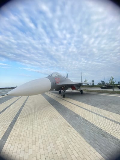 An exterior, low-angle, wide shot captures a fighter jet on display on a paved plaza under a cloudy sky. The jet is gray with red markings and has a long, pointed nose cone painted white. It is positioned on the right side of the frame, with its nose pointing towards the left. The plaza is paved with light-colored rectangular tiles arranged in a patterned walkway with dark gray stripes. In the background, a tank is visible on the right side of the frame, along with trees and streetlights. Two people, a man and a child, are standing near the jet. The sky is filled with a pattern of light and dark gray clouds.