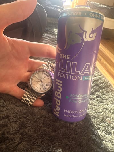 The scene, located in Höri, Switzerland, depicts a close-up indoor shot centered on a human hand, a wristwatch, and an energy drink can, resting on a dark grey, shaggy rug. A light-skinned human hand, with fingers partially obscuring the watch's bracelet, holds a silver-toned Rolex Oyster Perpetual Day-Date wristwatch. The watch features a fluted bezel, a jubilee bracelet, and a light-colored dial with Roman numerals, a date window displaying "12" at the 3 o'clock position, and a day window displaying "MONDAY" at 12 o'clock. The time indicated on the watch is approximately 10:10. Positioned immediately to the right of the hand and watch is an upright, purple Red Bull "The Lilac Edition" energy drink can. The can's label shows "OHNE ZUCKER" (sugar-free) at the top and "Waldmeister Pink Grapefruit" as the flavor, with "ENERGY DRINK Belebt Geist und Körper" at the bottom. The can exhibits a slight visible dent near its top rim. The background is softly blurred, revealing a dark fabric object (possibly a bed or couch) on the left and a light-colored floor on the right. Bright sunlight enters from an unseen source, casting distinct shadows and creating highlights on the rug and objects.