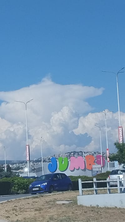 The image captures a bright, clear day in Pylaia Municipal Unit, Greece, with a striking blue sky dominating the upper portion of the frame. A prominent, large-scale sign displaying the word "JUMBO" in vibrant, distinct colors (blue, green, pink, orange, and purple) is visible. The sign is positioned in front of a building with a metallic facade.

In the foreground, a dark blue car is parked on the left side of the road, facing the sign. A portion of a silver car is visible on the right side of the frame, behind a white metal railing. The ground in the immediate foreground consists of dry, sparse vegetation and dirt.

Several tall streetlights with multiple arms are spaced along the roadway. Red and white banners are attached to two of the lampposts. In the background, a city skyline with buildings is spread across a hillside under a dramatic display of large, white cumulus clouds that create a sense of depth. Green foliage is visible on the left and right sides of the frame, partially obscuring the view of the surrounding area.