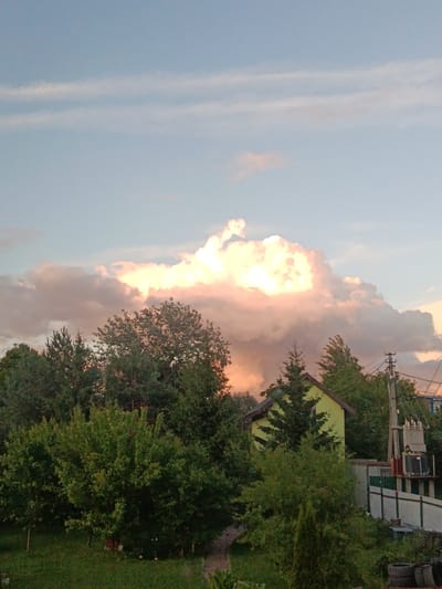 A wide-angle, slightly upward-looking shot captures a suburban landscape under a dynamic sky at dusk. The scene is dominated by a large, billowing cloud formation that is brilliantly illuminated from within by a warm, golden light, suggesting the setting sun. Wispy, pinkish clouds are scattered around this central mass, set against a pale blue sky.
Below the sky, a dense line of green trees forms a natural barrier across the middle ground. The trees vary in type, with some appearing as tall, dark evergreens and others as broader, leafy deciduous trees. Peeking through the foliage on the right side of the frame is the peak of a bright, lime-green house with a dark roof.
Further to the right, a utility pole with a large transformer and electrical wires stands tall, indicating the presence of infrastructure. A light-colored fence runs horizontally in front of the transformer. In the foreground, lush green grass covers the ground, with darker patches suggesting shadows. In the bottom right corner, a stack of dark tires is visible.
The overall impression is one of peaceful evening tranquility, with the dramatic cloud formation providing a striking visual focal point. The time of day is clearly evening, likely close to sunset, given the color of the sky and the illumination of the clouds. The setting appears to be a rural or semi-rural residential area, possibly in Motyakovo, Russia, based on the provided location context. No people are visible in the image. There is no visible text.